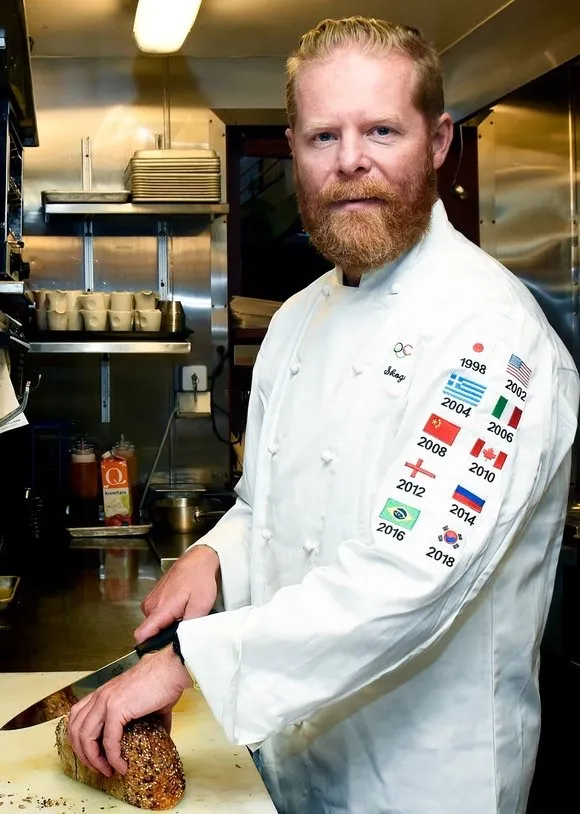 Trond Skogvoll i kokkejakke med flaggene fra flere olympiske leker, skjærer et nybakt frøbrød